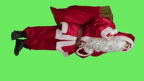 Vertical video Side view of santa claus character does ad against full body greenscreen backdrop, posing with red sack of presents to advertise christmas holiday season in studio. Saint nick