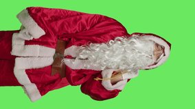 Father christmas shows mute symbol, espressing privacy and silence over greenscreen in studio. Santa claus with gifts bag doing hush sign to keep secret, private seasonal winter character.