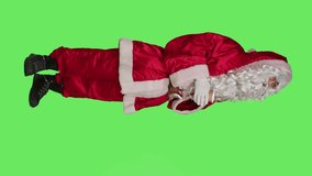 Vertical video Side view of santa claus drinks cup of hot chocolate in red festive winter costume with had and glasses, enjoying coffee beverage on greenscreen backdrop. Father christmas with seasonal