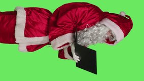 Vertical video Side view of santa claus man on online videocall waving at laptop screen to discuss with people about gifts, standing over greenscreen background. Person in red festive suit on