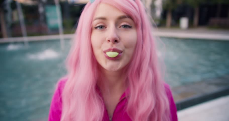 Cheerful infantile girl in pink inflates a yellow ball of gum while sitting near a fountain outdoors in summer Royalty-Free Stock Footage #1106055749