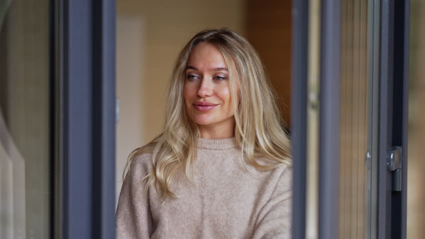 Beautiful smiling woman standing at glass door waving inviting inside looking away. Positive charming Caucasian lady on porch at home indoors