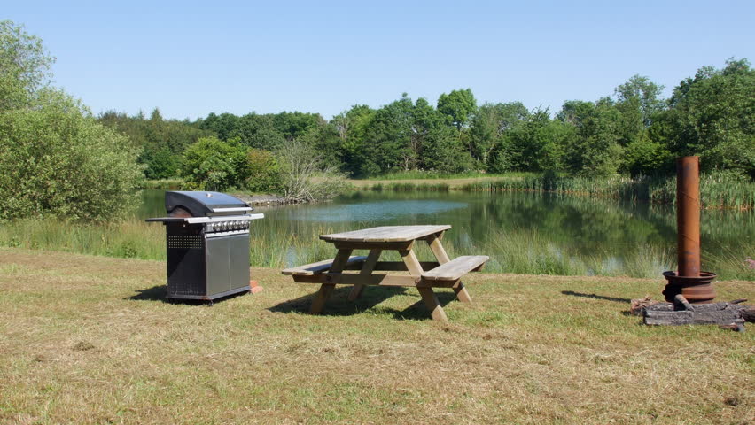 BBQ and picnic wooden bench next to a beautiful lake. A metal gas fired barbecue on the grass bank.