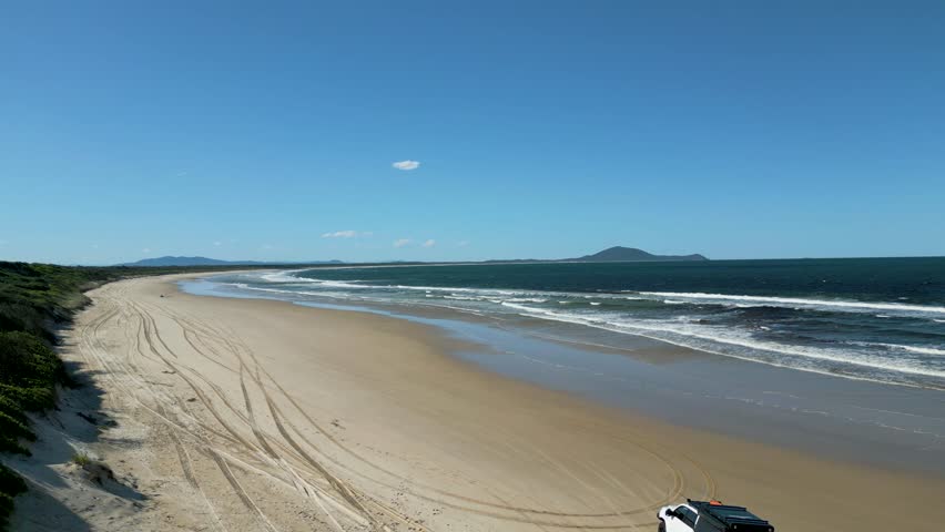 Aerial view of a Single Car driving along a long sandy beach. a beautiful blue sky day with the waves rolling in.