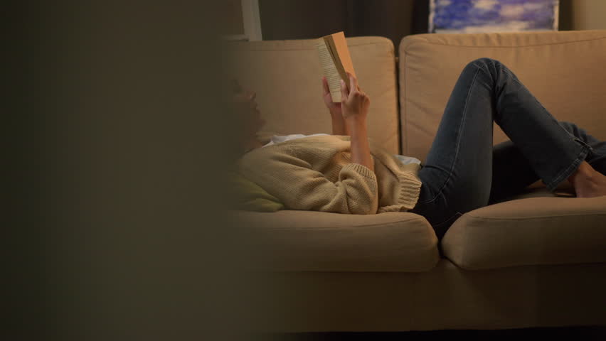 Happy woman opening, reading and relaxing a book on relax time at home. Vacation time of female reading a book on sofa at night. Education concept. Royalty-Free Stock Footage #1106070949