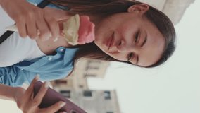 Vertical video, Close up, beautiful girl with long brown hair, dressed in blue shirt, enjoys ice cream and looks through photos, videos on mobile phone while standing on square of old city