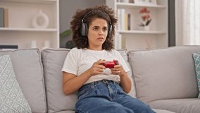 Young beautiful hispanic woman playing video game sitting on sofa at home