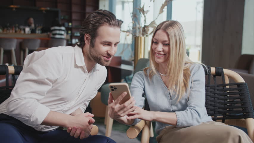 Attractive young happy couple with a smartphone in a cafe. Talking smiling sitting modern interior. Have conversations together. Woman and man. Slow motion | Shutterstock HD Video #1106090305
