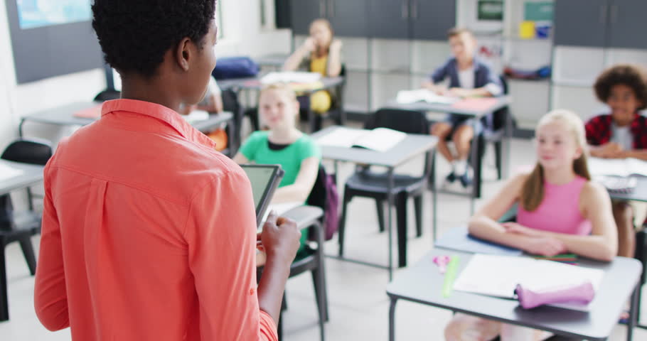 Portrait of diverse female teacher and happy schoolchildren at desks in school classroom. Education, inclusivity, elementary school and learning concept.