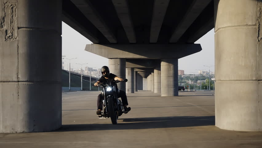 Biker riding a motorcycle on a summer evening. Black chrome motorcycle. Protective helmet. Fast driving. Place under the road bridge. Shadow on the asphalt Asphalt parking. Protective helmet on a man.