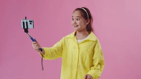 Portrait of a smart asian girl kid wearing yellow rain coat, holding a selfie stick with phone in her hands and posing, doing selfie on colourful background wall in studio