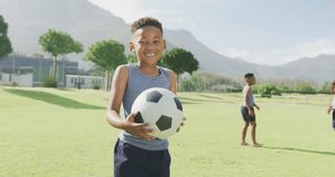 Video portrait of smiling african american schoolboy holding football in field. Education, childhood, inclusivity, health, sport, elementary school and learning concept.