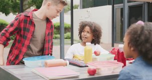 Video of three diverse schoolchildren talking at lunch sitting in schoolyard. Education, childhood, inclusivity, elementary school and learning concept.