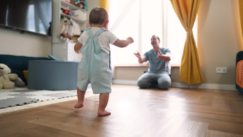 Baby's first steps. Father helps encourages Toddler to take bold first steps at home. Parent lends helping hand for teaches of baby. Toddler walk at home. Baby first active steps on father's day Royalty-Free Stock Footage #1106102589