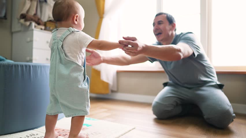 Baby's first steps. Father helps encourages Toddler to take bold first steps at home. Parent lends helping hand for teaches of baby. Toddler walk at home. Baby first active steps on father's day | Shutterstock HD Video #1106102589