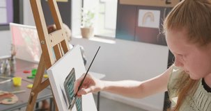 Video of happy caucasian schoolgirl concentrating at easel, painting in art class, copy space. Education, childhood, creativity, inclusivity, elementary school and learning concept.