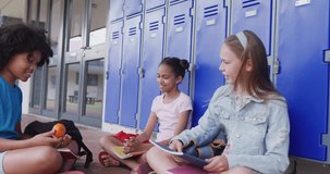 Video of three happy, diverse schoolgirls talking, sitting by lockers in school corridor, copy space. Education, childhood, inclusivity, elementary school and learning concept.