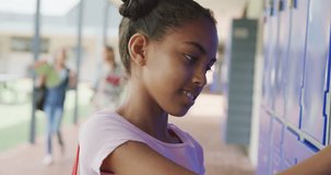 Video portrait of happy biracial schoolgirl smiling by lockers in school corridor, copy space. Education, childhood, inclusivity, elementary school and learning concept.