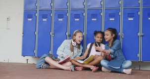 Video of three happy, diverse schoolgirls looking at smartphone, sitting in corridor, copy space. Education, childhood, inclusivity, elementary school and learning concept.
