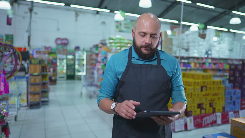 Brazilian employee of Grocery store using tablet device checking inventory standing inside supermarket chain,w wearing apron and engaged with work Royalty-Free Stock Footage #1106104247