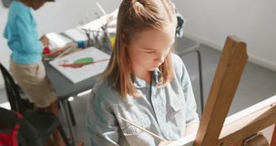 High angle video of concentrating caucasian schoolgirl at easel painting in art class, copy space. Education, childhood, inclusivity, creativity, elementary school and learning concept.