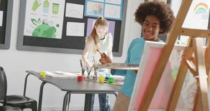 Video of happy diverse girl and boy talking by boy's easel in art class, copy space. Education, childhood, inclusivity, creativity, elementary school and learning concept.