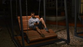 Man making a video call while sitting on wooden swing. Media. Young man using smartphone for calling late in the evening outdoors.