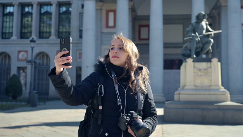 Pensive young Latin woman in warm clothes with professional photo camera taking selfie on smartphone while standing near Prado Museum in Madrid, Spain