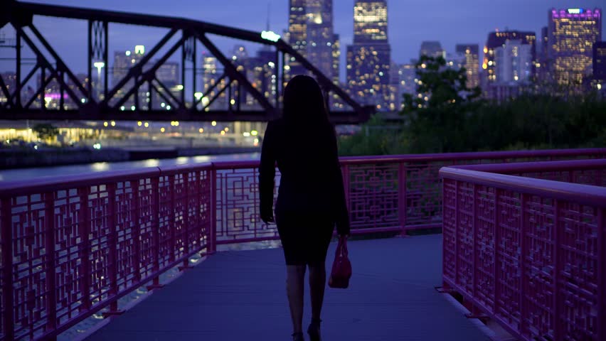 African American business woman walking along a bridge at night wearing a fashionable business black dress. The Chicago building skyline is illuminating the river and park as she confidently walks
