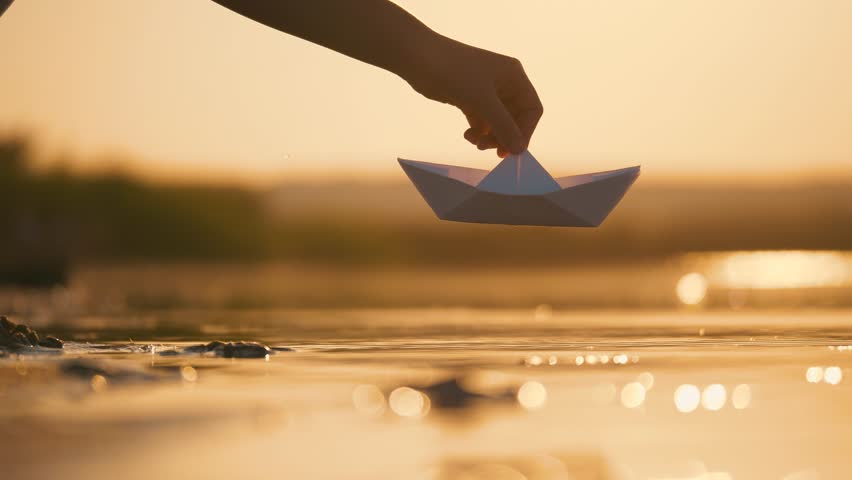 A woman's hand launches white paper craft on water at beach. Paper boat on water. Close-up of girl's hand with boat. Floating paper boat on water. Royalty-Free Stock Footage #1106132339