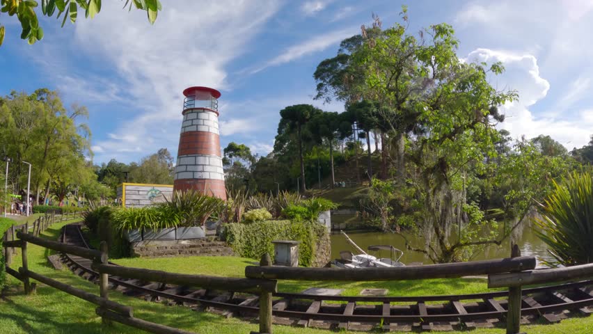 Time lapse of towering lighthouse and train ride in natural park with lake, trees, ecological trails, beautiful clouds and blue sky	