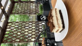 Eating delicious tiramisu dessert cake at a wooden table on a city cafe terrace. Taking a piece of cake with a dessert fork. Lifestyle vertical video.