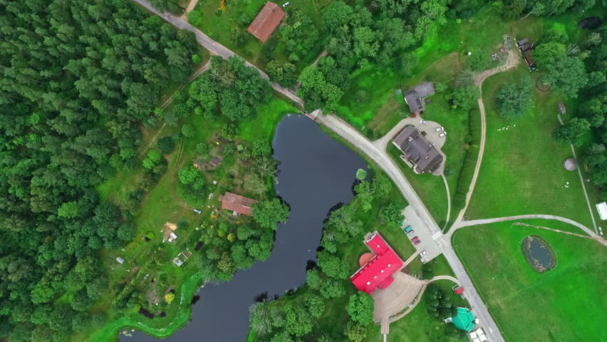 Drone view tilting downward of a rural community surrounded by trees with a small lake in the center. Few homes, buildings, outbuildings and roads throughout.