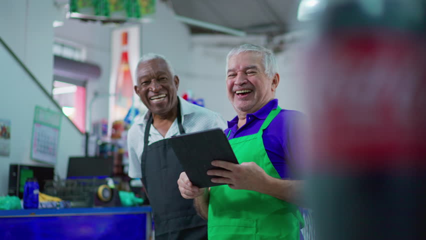 Joyful diverse Brazilian senior staff workers of supermarket chain smiling at camera with table and uniforms. African American older employee and a caucasian person laughing together Royalty-Free Stock Footage #1106149619