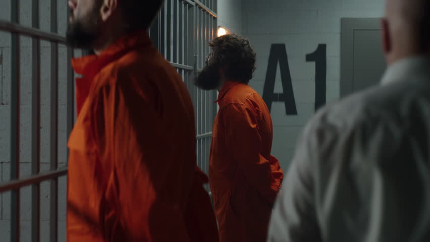 Two prisoners in orange uniforms stand facing the metal bars in front of prison cells. Prison officer with police baton walks, watches criminals in jail. Detention center or correctional facility. Royalty-Free Stock Footage #1106150193