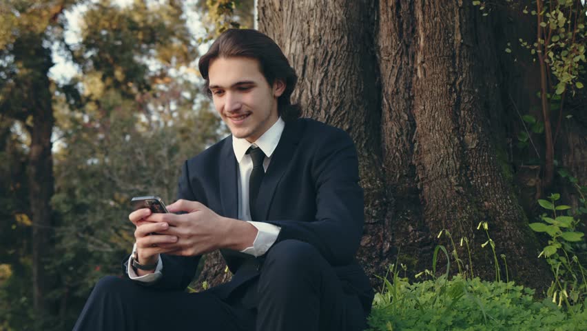 Smiling businessman in the city  looking to a cell phone. Confident and happy man in a business suit is sitting outdoors and using  smartphone. Young man in a suit with mobile phone, slow motion. 