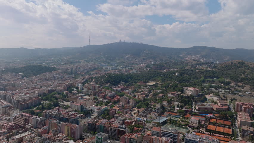 Fly high above metropolis. Aerial panoramic view of large city in hilly landscape. Silhouette of tall TV tower and Temple of the Sacred Heart of Jesus on hill above city. Barcelona, Spain