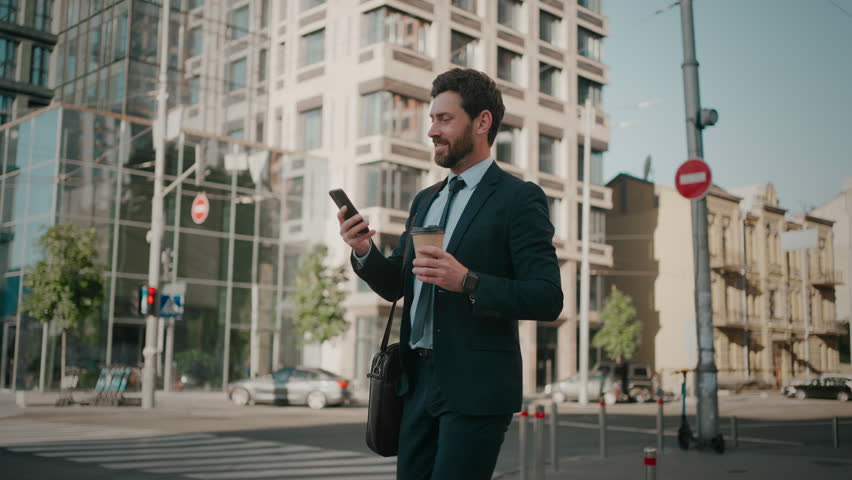 Smiling bearded businessman goes to work chatting on social networks on phone. Man drinks coffee from disposable cup walking down city street in morning low angle shot slow motion Royalty-Free Stock Footage #1106158203