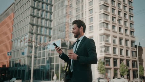 Smiling bearded businessman goes to work chatting on social networks on phone. Man drinks coffee from disposable cup walking down city street in morning low angle shot slow motion Arkivvideo