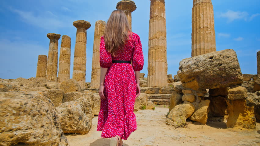 A female tourist visits the antique Temple of Hercules in Sicily, Italy. A young woman in a beautiful long dress enjoys the view of the greek majestic temple with massive columns in Agrigento. Royalty-Free Stock Footage #1106164565