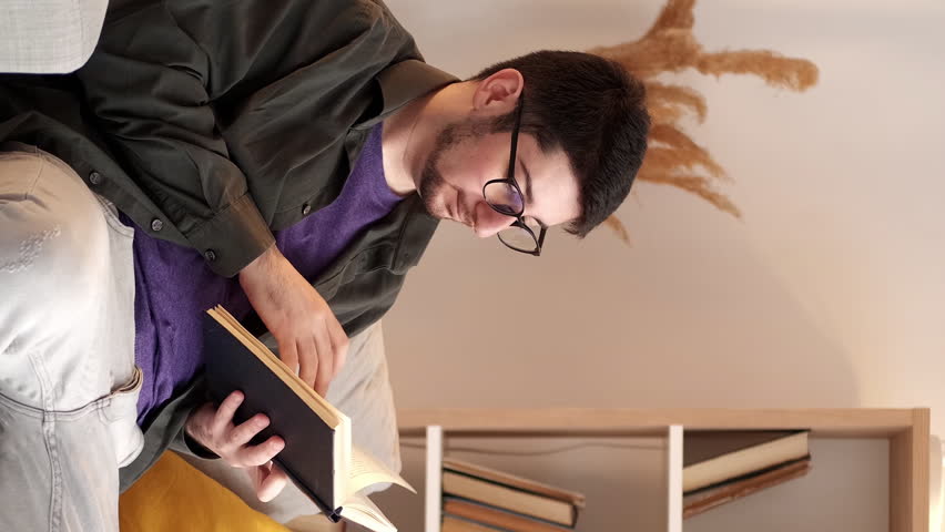 Vertical video. Home reading. Book leisure. Weekend relaxing. Peaceful man in glasses enjoying literature resting on comfortable sofa in cozy living room.