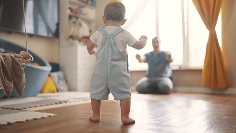 baby first steps. baby goes her father at window learns to walk to take first steps. happy family kid dream concept. dad calls son baby first steps indoors. happy family indoors concept lifestyle วิดีโอสต็อก