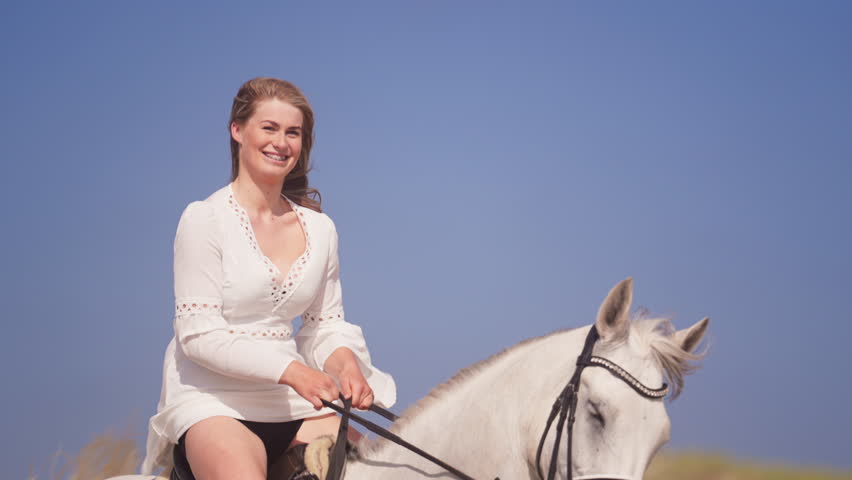 Medium shot of a long-haired girl smiling with braces riding a white horse during a sunny day