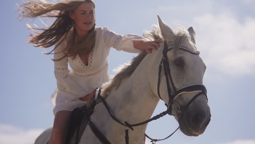 A medium shot of a girl wearing a white dress, petting her white horse during a windy day