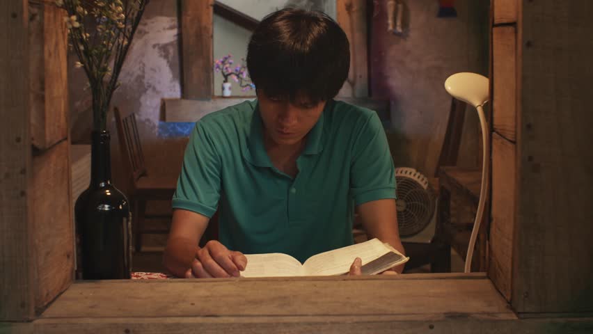 Looking through an open window at a young Asian male reads a book at a desk in a rustic room. Locked tele shot Royalty-Free Stock Footage #1106190043