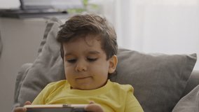 Front view small kid boy enjoy using modern gadget smart phone looking at mobile screen, watching cartoons, having fun, playing mobile online games, sit on couch at home. Close up