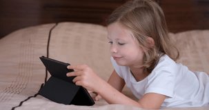 Cute little girl lying on the sofa looks at the screen of the tablet