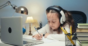 Little Girl in Headphones Learns Language Online, Uses a Laptop, Looks at the Screen, Performs School Assignments at Home, Writing Notes, Listening to Lectures or Music, Distance Learning