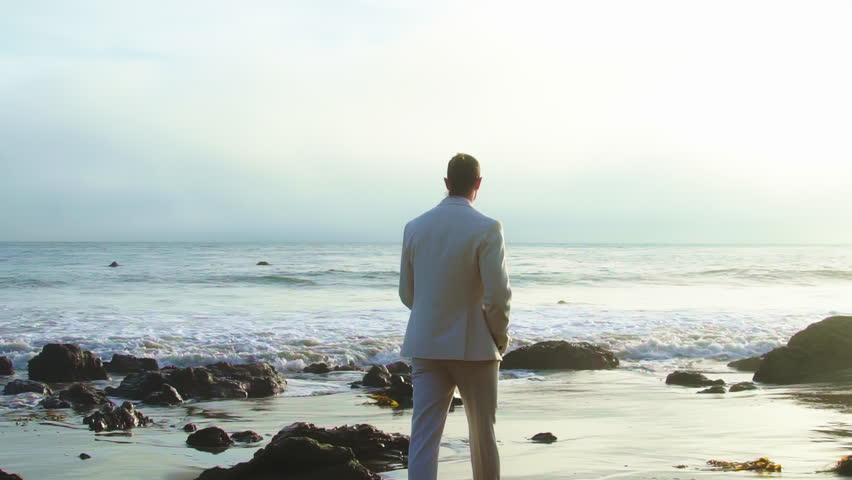 Handsome man in white suit walking by Pacific Ocean. Man wearing stylish outfit on the beach. Back view Royalty-Free Stock Footage #1106217003