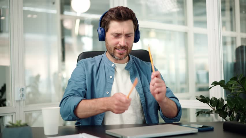 Happy funny business man in blue headphones dancing at workplace celebrating good news pretend playing drums with pencils having fun enjoying work break listen music singing song at office. Royalty-Free Stock Footage #1106219303